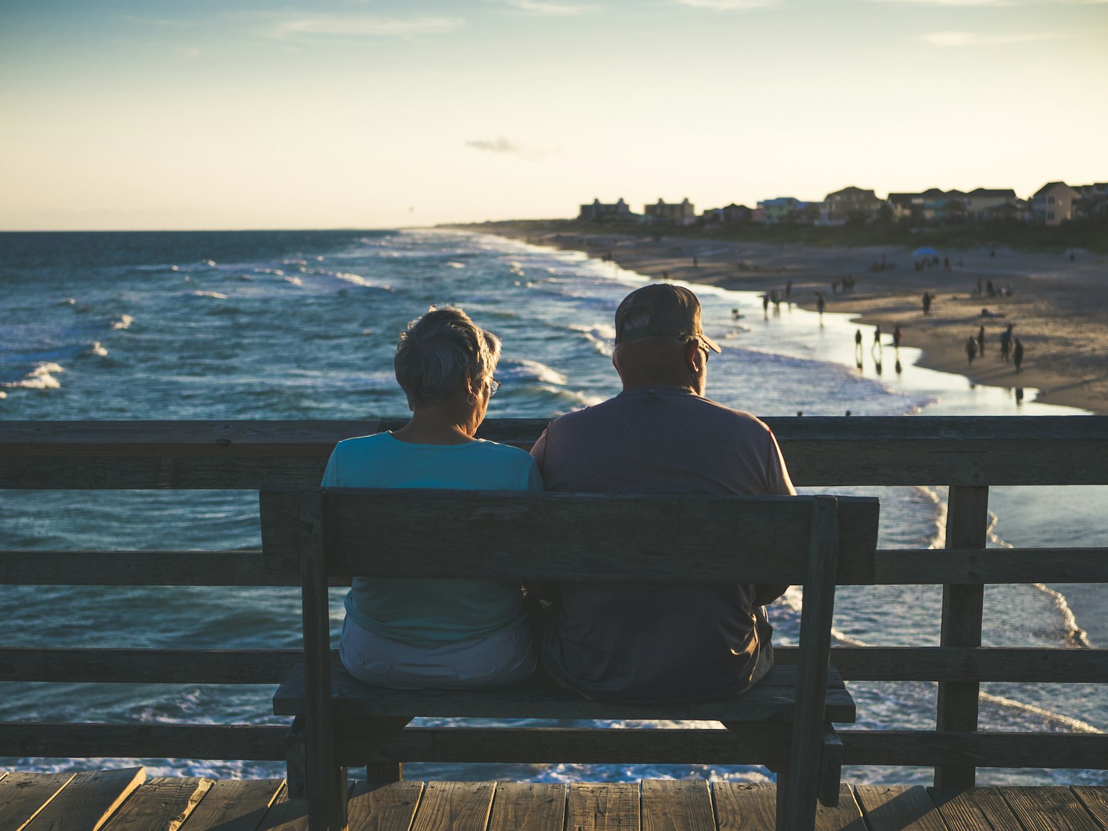 An older couple seen from behind, sitting together on a bench above the shore.