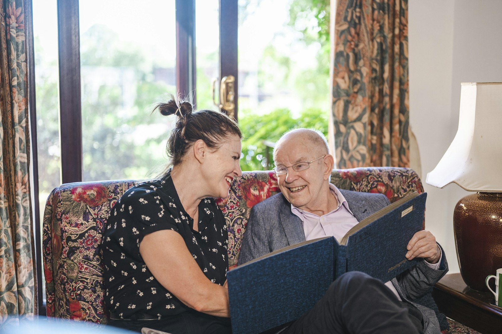 An adult daughter and her older father reading a large bound record together on a sofa.