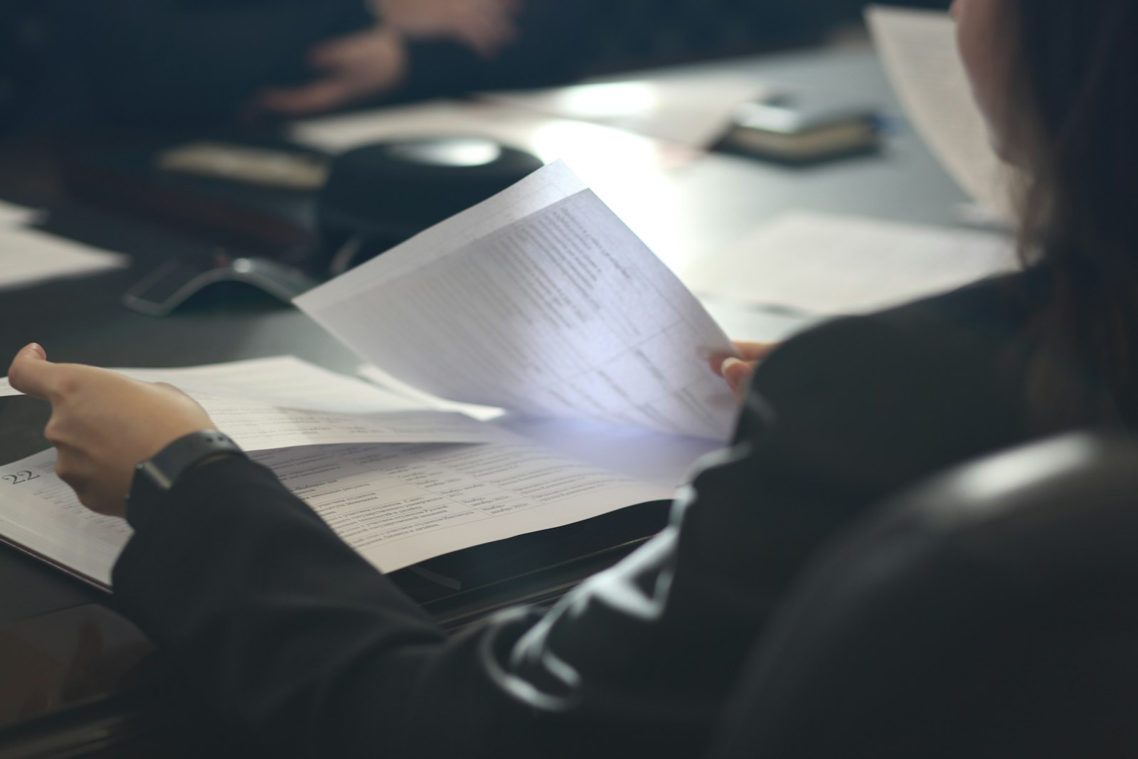 A person at a meeting table reading through a sheaf of printed documents.